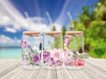Drei 16 oz Glasbecher mit Bambusdeckel und Glastrinkhalm, verziert mit zarten Blumen- und Schmetterlingsmotiven in Rosa- und Violetttönen. Die Becher stehen auf einem Holztisch vor einem Hintergrund mit Strand und Palmen, was eine frische, sommerliche Atm